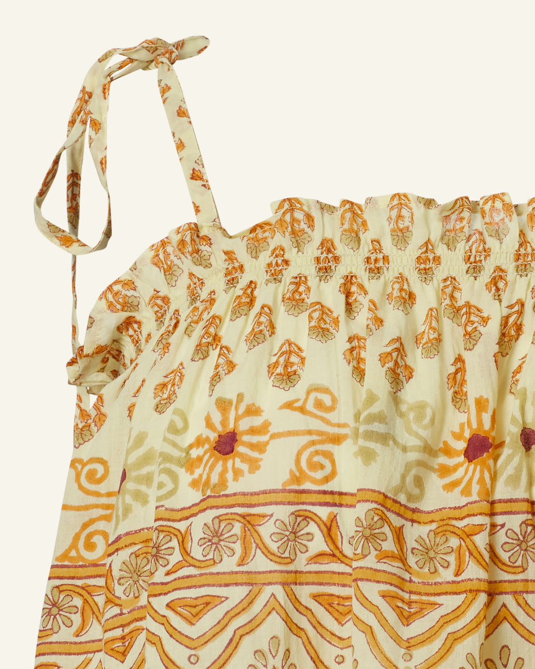 A close-up of the Lila Saffron Bhutti Top reveals its sleeveless cotton voile design, featuring a gathered neckline. The fabric displays an intricate floral and geometric pattern in vibrant shades of orange, yellow, and beige. These patterns are enriched with block-printed details and completed with a tie strap on the shoulder.