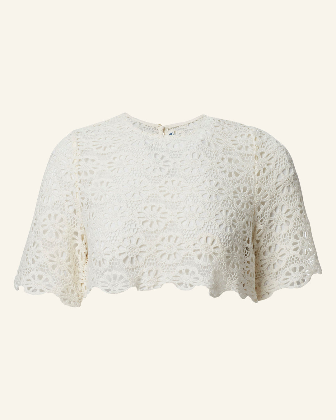 The Nala Daisy Lace Cropped Top is a white, cotton crochet top featuring daisy floral patterns on a light beige background. It has short sleeves, a scalloped hem, a round neckline, and a button closure at the back.