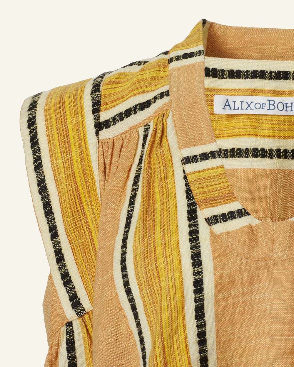 Close-up of the Nehru Sun Stripe Fringe Top featuring a label reading "ALIXOFBOHEMIA." This striped sleeveless garment showcases vertical yellow, black, and tan hues in hand-loomed cotton, accented with pleated detailing near the shoulder.