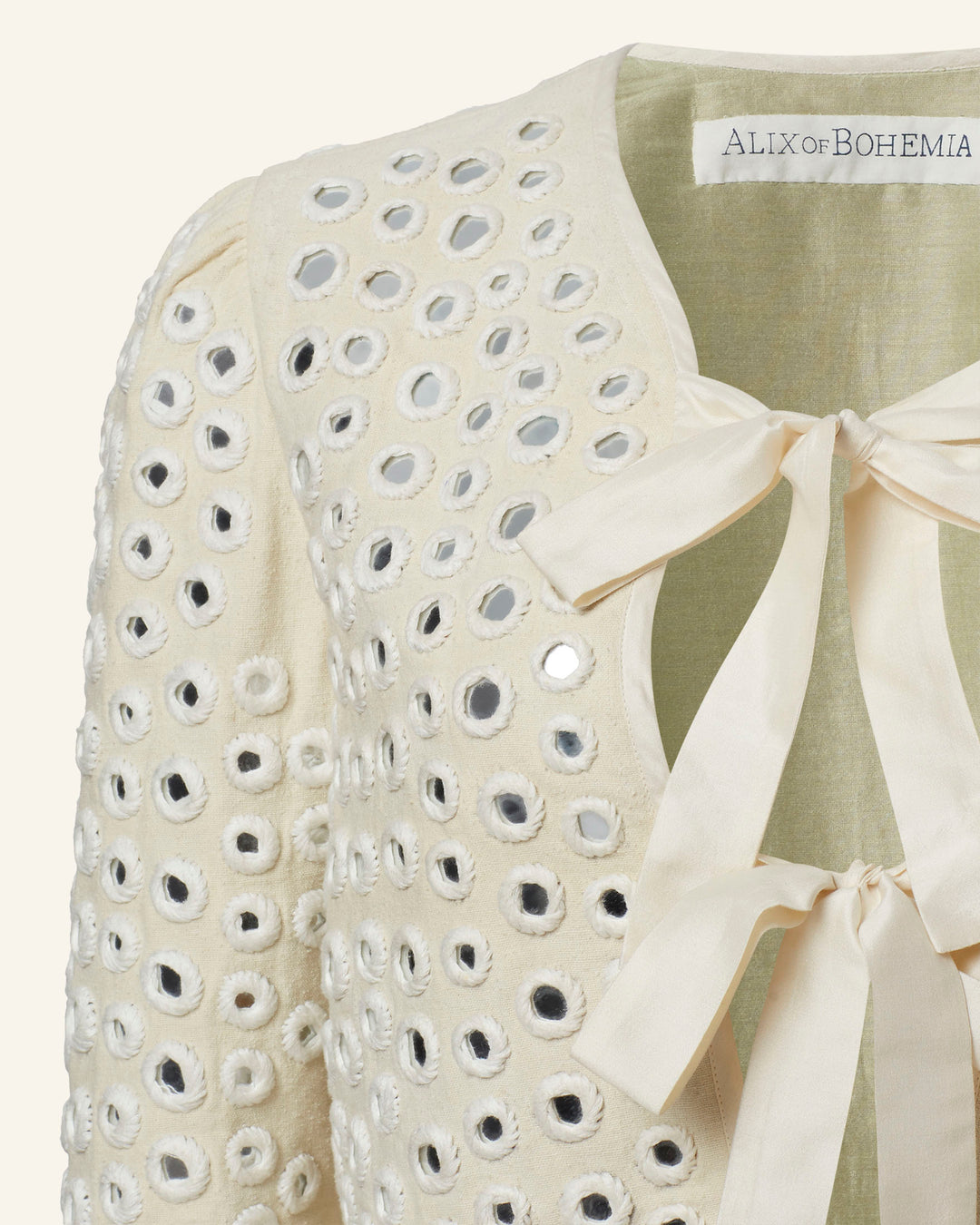 Close-up of the Pasha Frosted Mirror Jacket in cream-colored silk, featuring circular cut-out details with intricate mirror work and two cream bows at the neckline. A label reading "ALIX of BOHEMIA" is visible at the top.