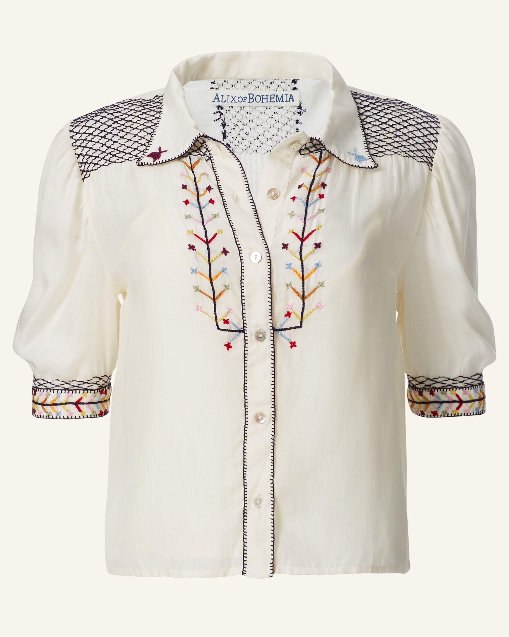 Introducing the Peyton Aurora Arrow Blouse, a Tencel blouse in cream with short sleeves. It features intricate hand-embroidered geometric and floral patterns adorning the shoulders, front, and cuffs. This folk-inspired piece is completed with buttons down the front and a collar.