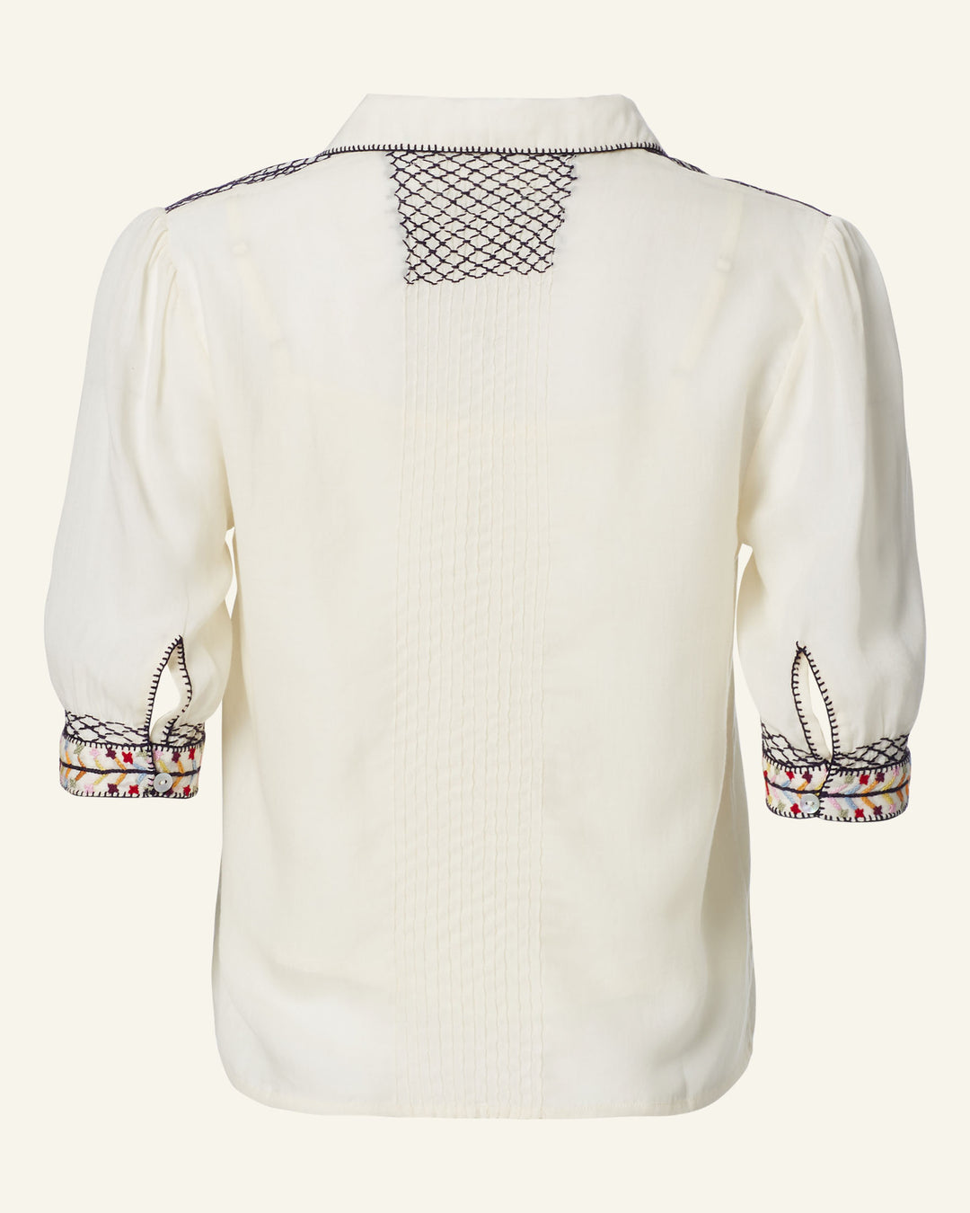 Discover the Peyton Aurora Arrow Blouse, a white folk-inspired top featuring intricate black stitching on the shoulders and back. Its hand-embroidered three-quarter sleeves boast patterned cuffs that add vibrant accents. The blouse is crafted from a lightweight, slightly translucent fabric with textured vertical lines.