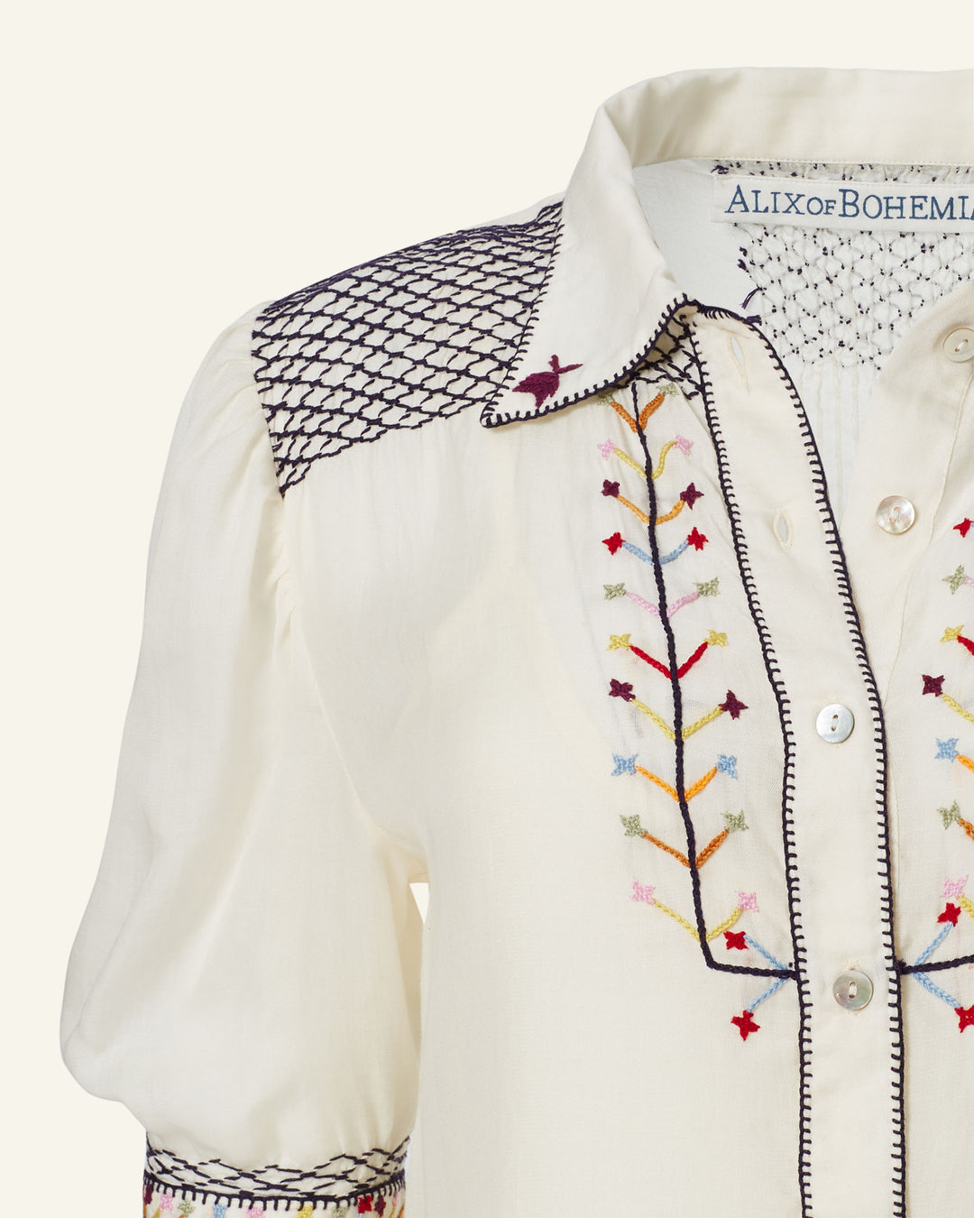 Close-up of the Peyton Aurora Arrow Blouse, showcasing its cream color and intricate hand-embroidered details in various colors. This folk-inspired design features puffed sleeves, visible buttons, a diamond pattern on the shoulders, and small floral designs down the front. The label "ALIX of BOHEMIA" is displayed inside.