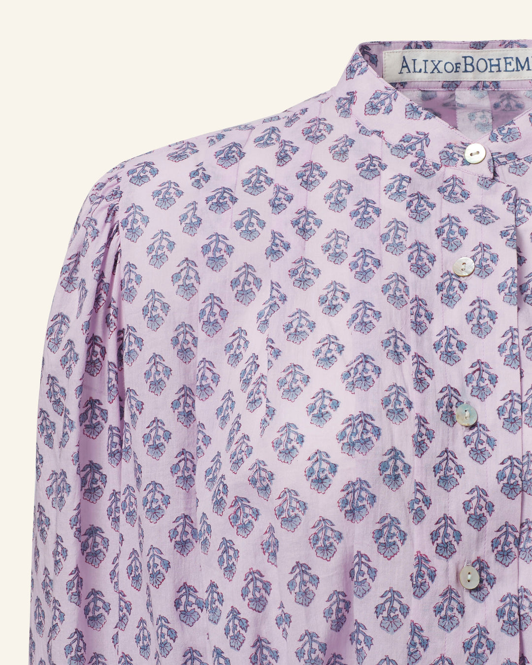 The Ryland Lilac Bhutti Top displays a detailed view of a lavender floral print in light purple, highlighted with blue and red leaf patterns. Made from cotton voile, this blouse features a Victorian silhouette with a high collar and button-down front. Inside the collar, a fabric label reads "ALIX of BOHEMIA.