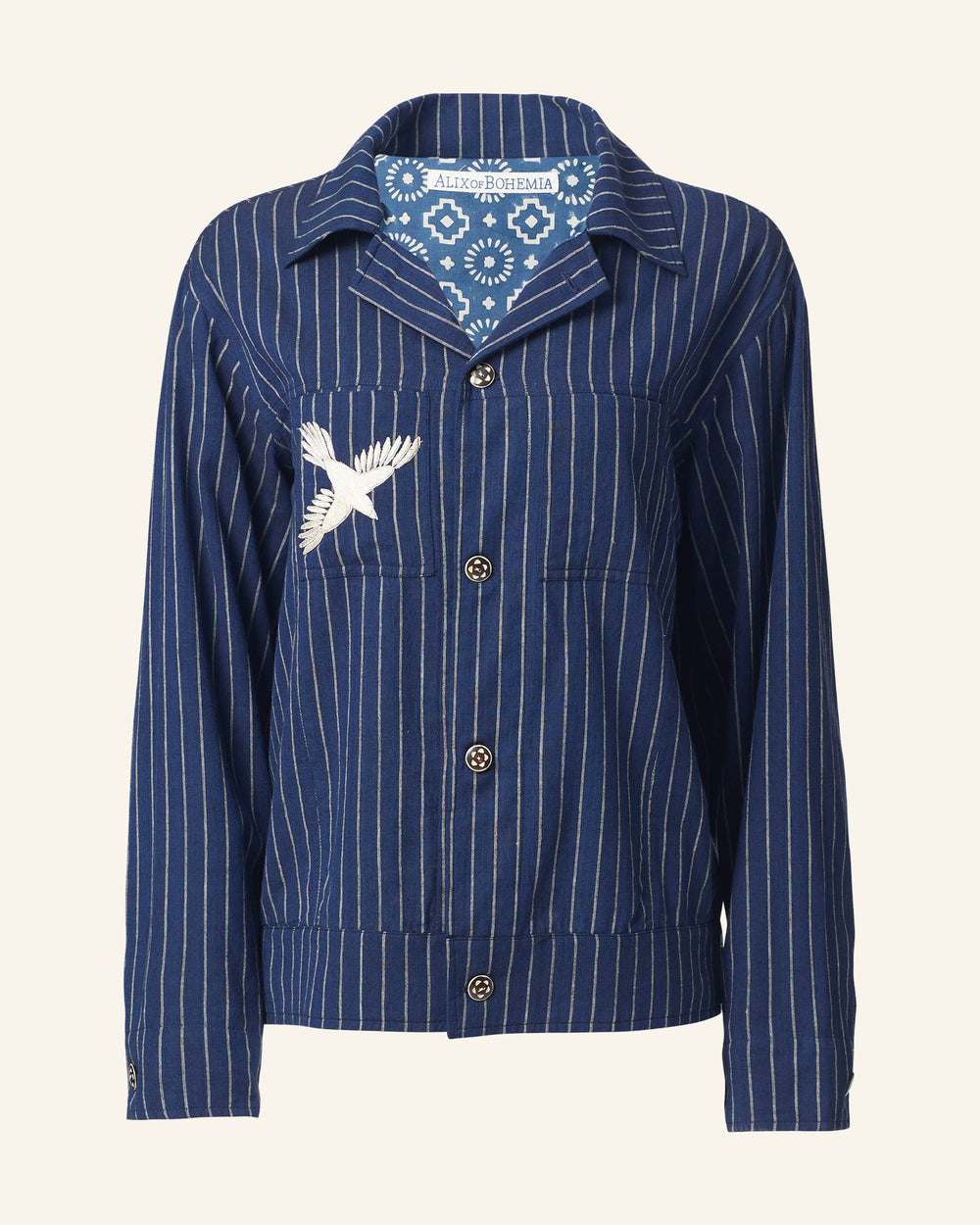 The Seba Luna Night Jacket is a blue, vertically striped button-up crafted from Khadi cotton, adorned with hand-embroidered white bird artwork on the left chest pocket. It features a pointed collar and is finished with dark buttons that have a circular design, making it a striking piece.