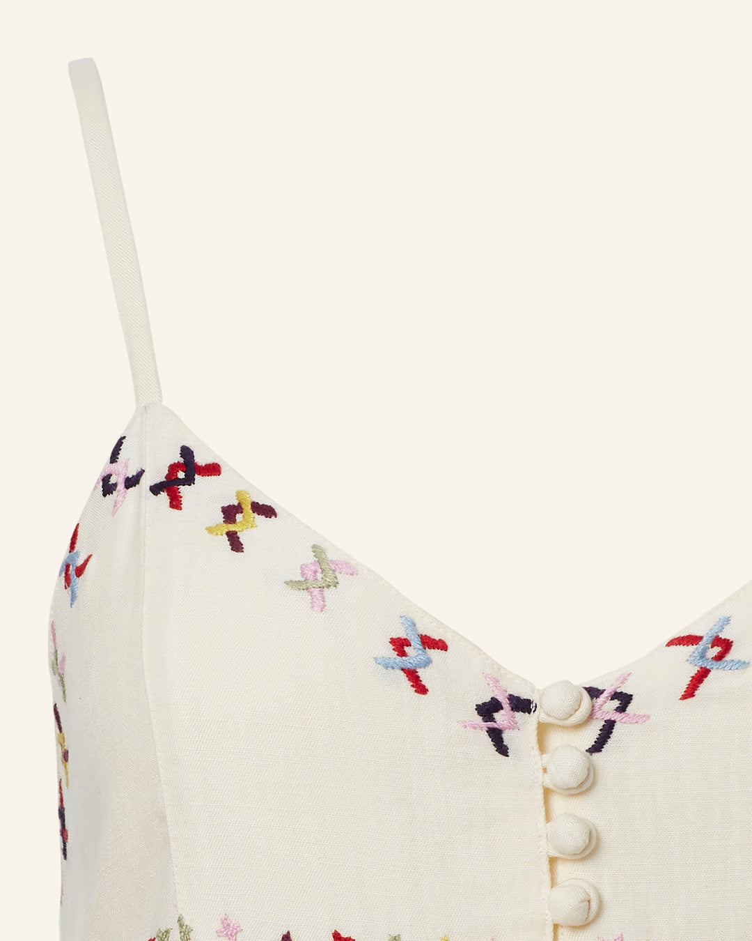 This close-up showcases the Skylar Aurora Arrow Dress, featuring a hand-embroidered, cream-colored top with colorful cross-stitch details and fabric-covered buttons. Thin spaghetti straps accentuate the design, highlighting vibrant patterns in red, purple, blue, and yellow. Perfect for pairing with your favorite summer ensemble.