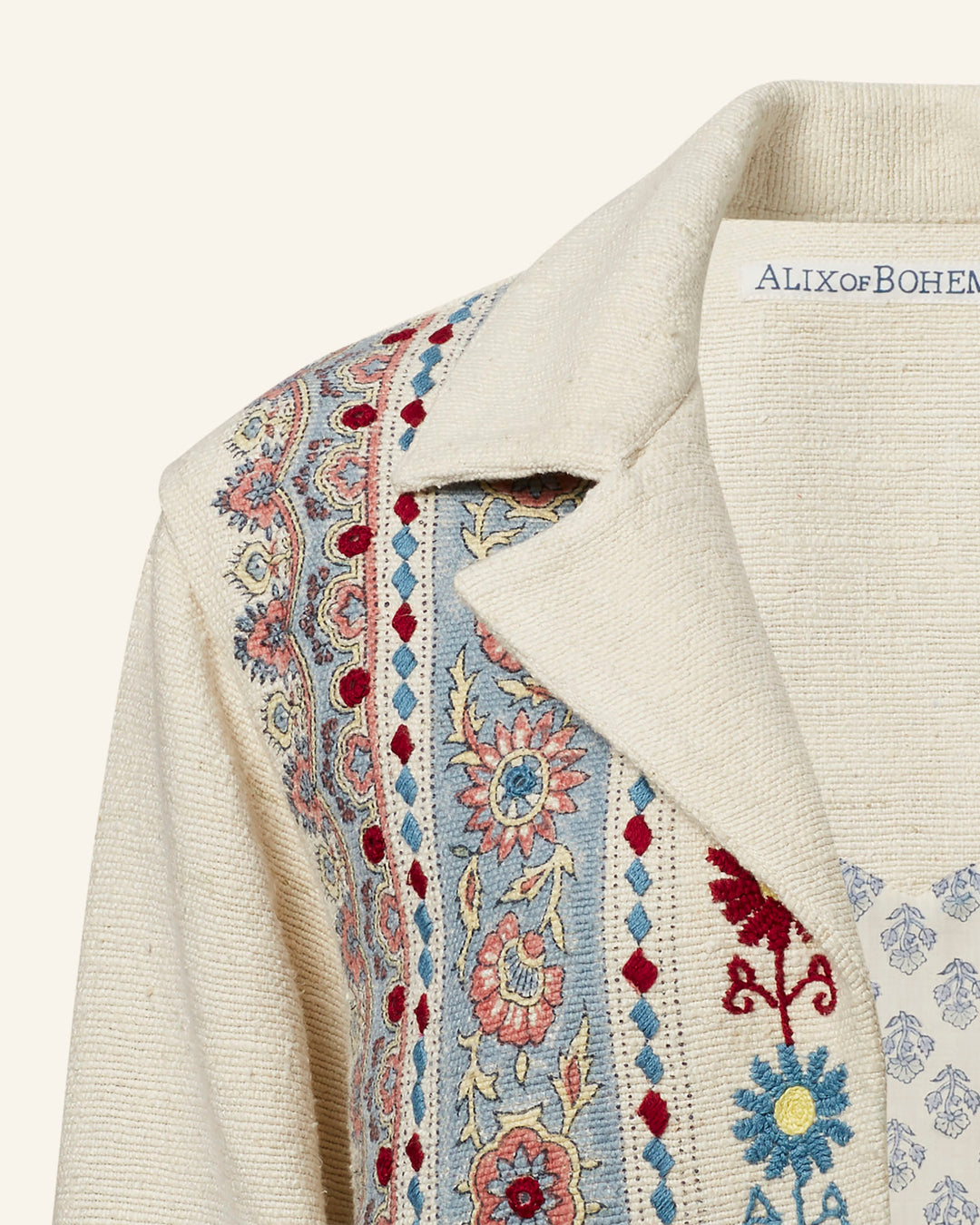 A close-up of the Western Riviera Silk Jacket showcases its intricate hand-embroidered details, featuring floral and geometric patterns in red, blue, and gold tones on beige silk. The "Alix of Bohemia" label is visible on the inside collar. This design exudes a bohemian style, making it a stunning statement piece.