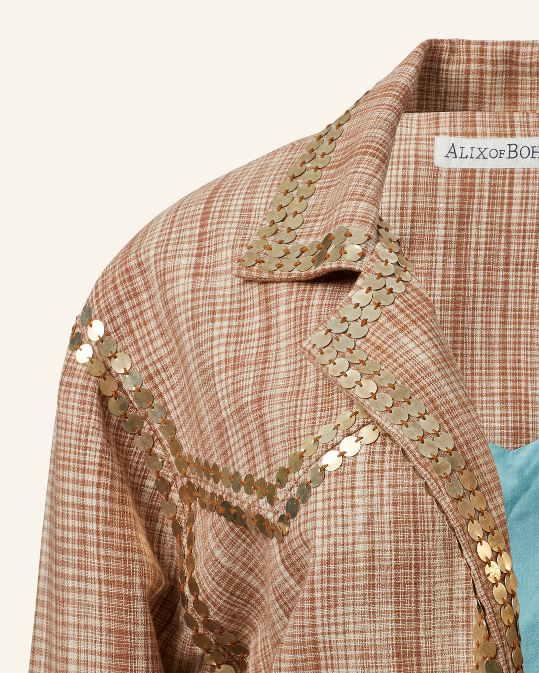 A close-up of the Western Sand Coin Jacket reveals a brown and cream checkered garment made from khadi cotton, highlighting the collar and shoulder detail. The hand-embellished gold sequins arranged in a V-shaped pattern add an artisanal touch. A small white label with text is partially visible at the top.