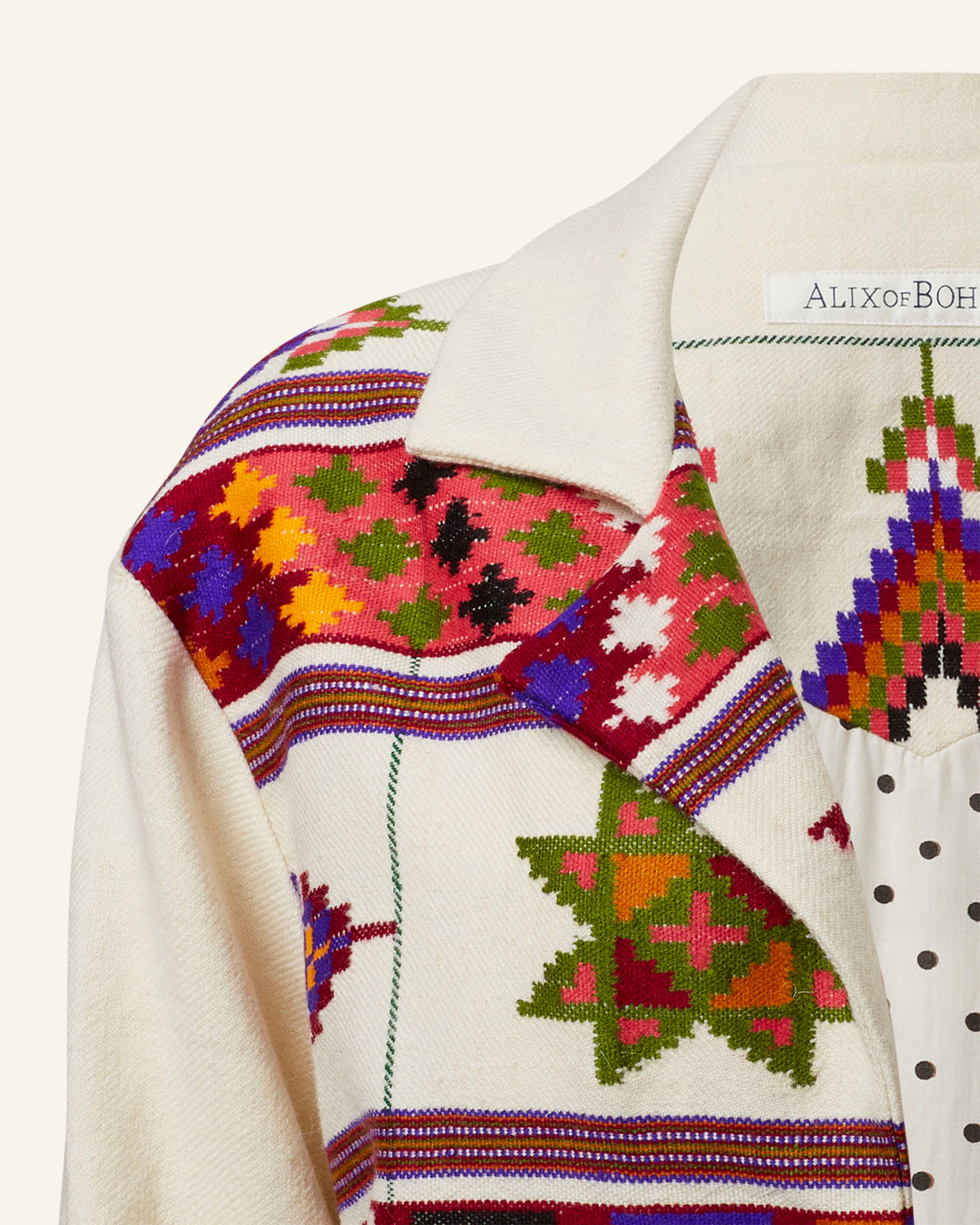 A detailed view of the Western Tiger Eye Blanket Jacket showcases its vibrant geometric star patterns in red, green, purple, and blue hues set against a cream background. Crafted from hand-woven Kullu wool fabric, the piece bears the "Alix of Bohemia" label.