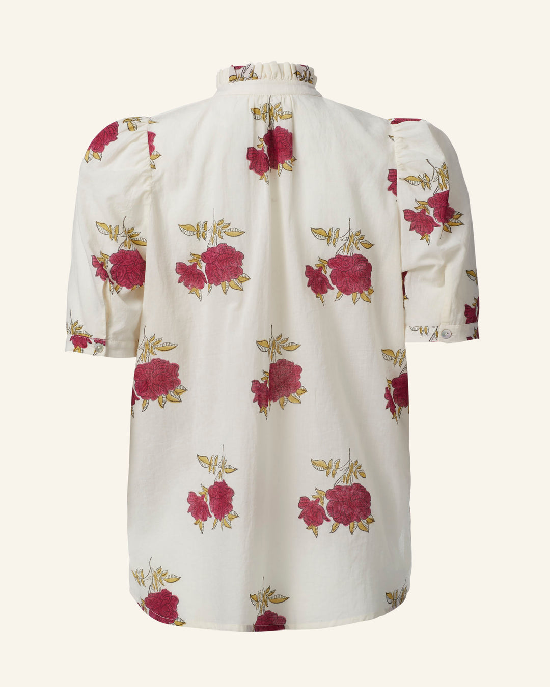 The Winnie Cerise Rosette Shirt is a cream-colored blouse made from cotton voile, featuring a high collar and short puff sleeves. The hand-block printed pattern showcases large red roses with green leaves, and the fabric has a textured appearance. This rose motif shirt is set against a plain background.