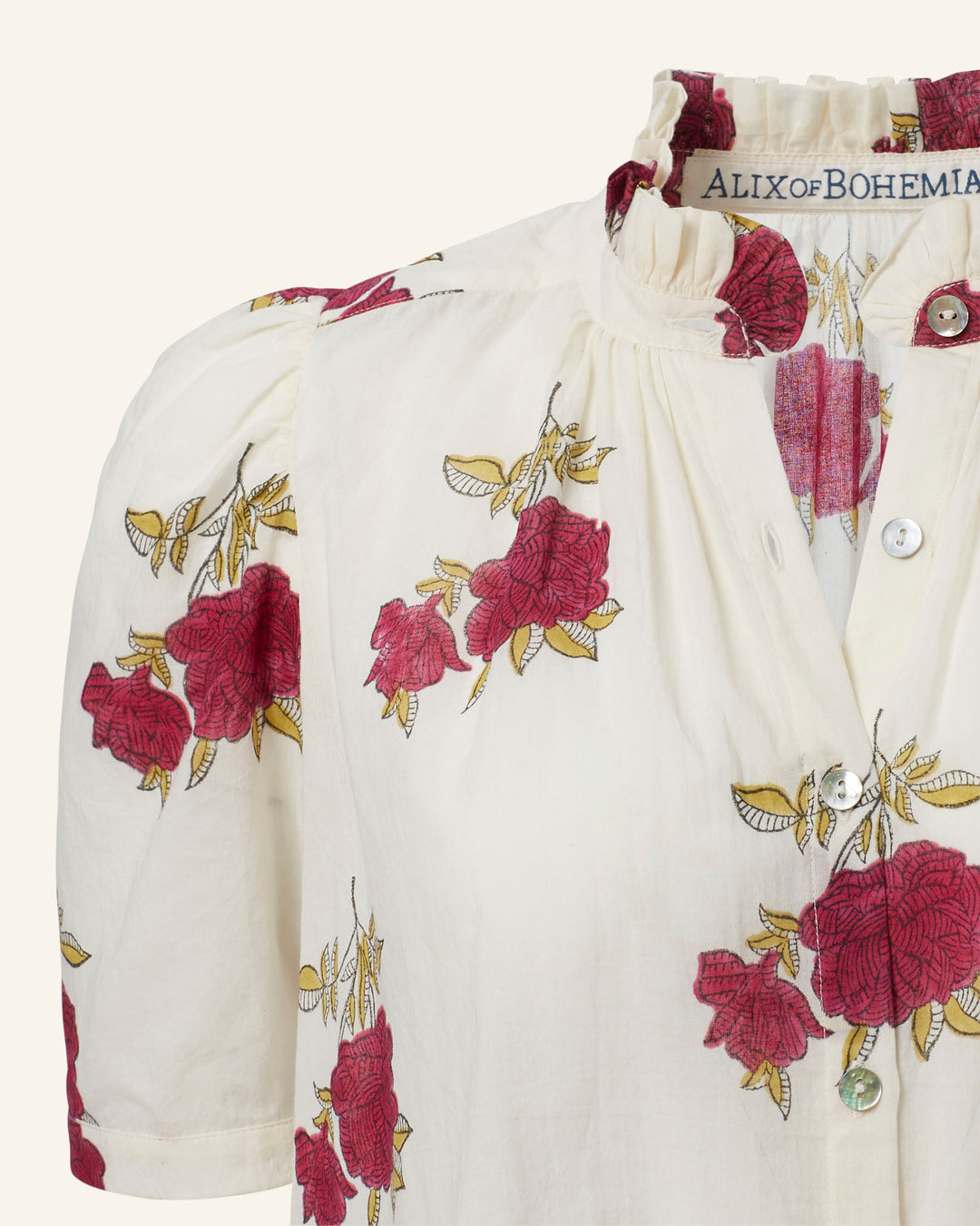 Close-up of the Winnie Cerise Rosette Shirt in cotton voile, showcasing a floral pattern with red roses and green leaves. The blouse features a ruffled neckline and buttons down the front, with the label "Alix of Bohemia" highlighting its hand-block printed elegance.