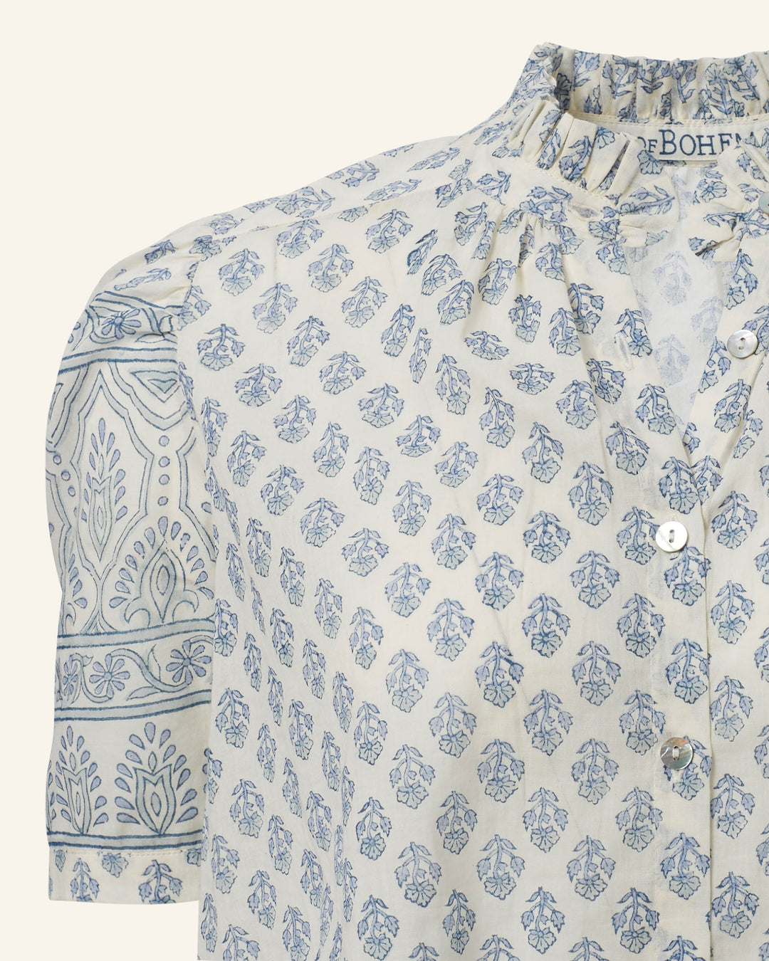 A close-up of the Winnie Delft Bhutti Shirt reveals its vintage-inspired design, showcasing a light cream cotton voile blouse with blue leaf and floral patterns. It features a ruffled collar, short sleeves adorned with intricate designs, and button details down the front.