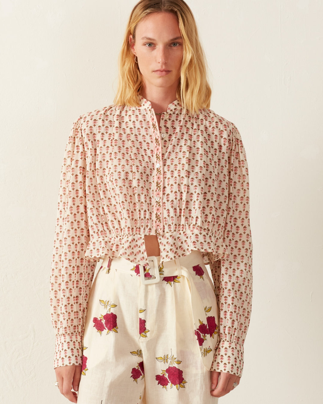 A person with long, blonde hair stands against a neutral background, wearing the Ryland Ruby Quatrefoil Top. This blouse is crafted from cotton voile and features long sleeves and a button-down front. It's cinched at the waist with a matching belt for an elegant look, paired beautifully with high-waisted pants adorned with large red flowers.