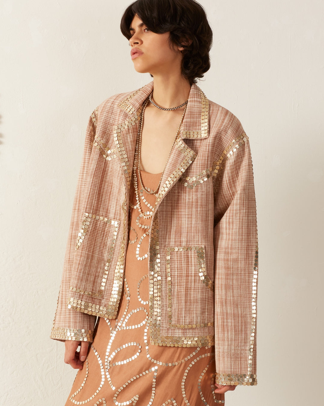 A person with short dark hair models a stylish outfit featuring the Western Sand Coin Jacket, a peach-colored plaid jacket adorned with meticulously hand-embellished gold sequins. Underneath, they wear a patterned dress, set against a simple white wall backdrop.