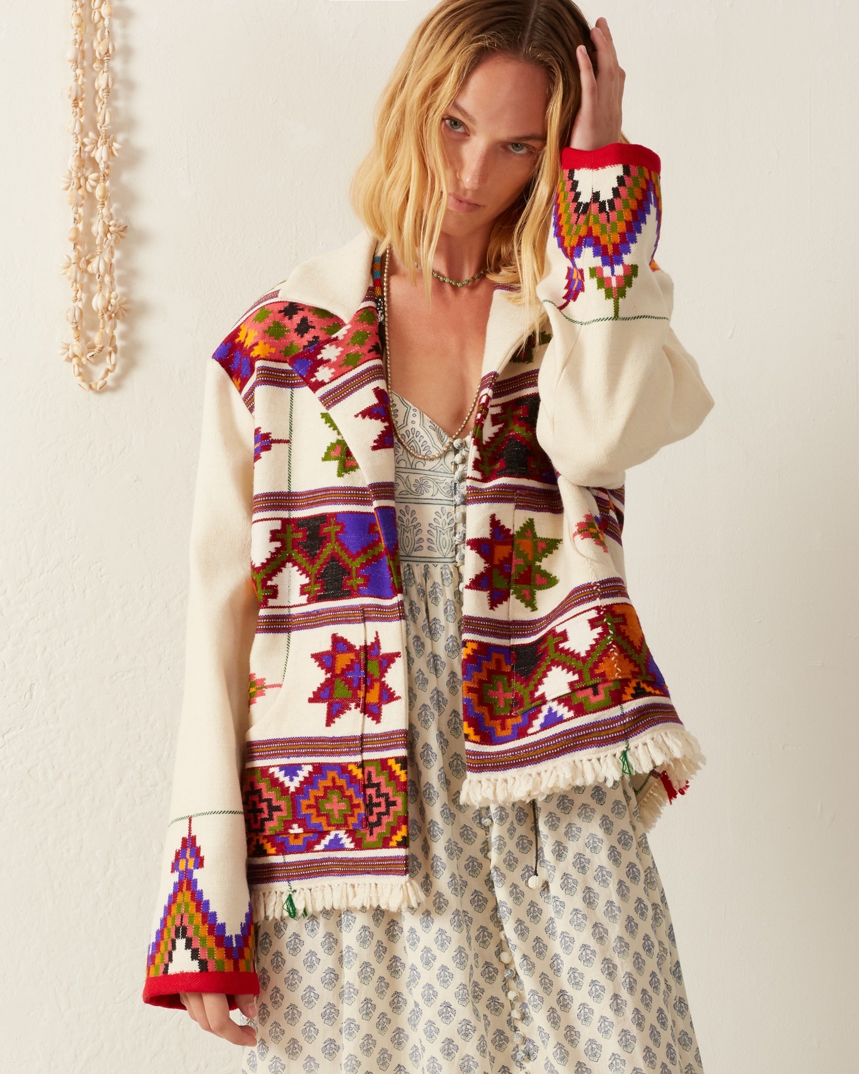 A person stands against a light background wearing the Western Tiger Eye Blanket Jacket, a cream-colored, Western-inspired statement piece adorned with colorful geometric patterns and fringe on the edges, over a patterned dress. They have one hand raised to their head and are looking towards the camera.