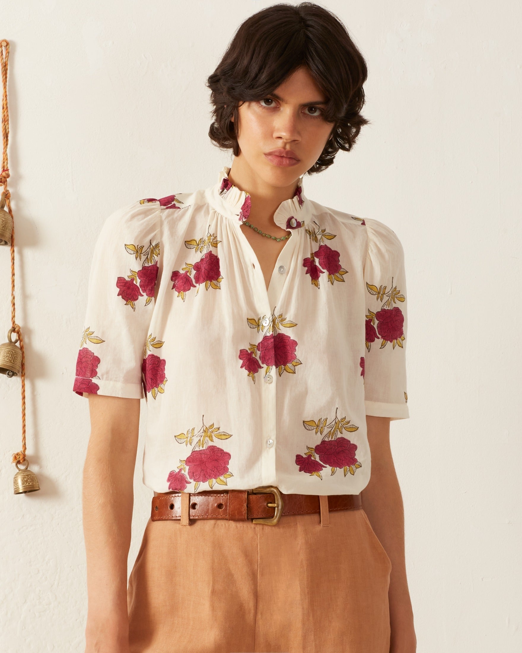 A person wearing the Winnie Cerise Rosette Shirt and beige high-waisted pants, cinched with a brown belt. Their short dark hair complements the light background adorned with hanging bells on the left.