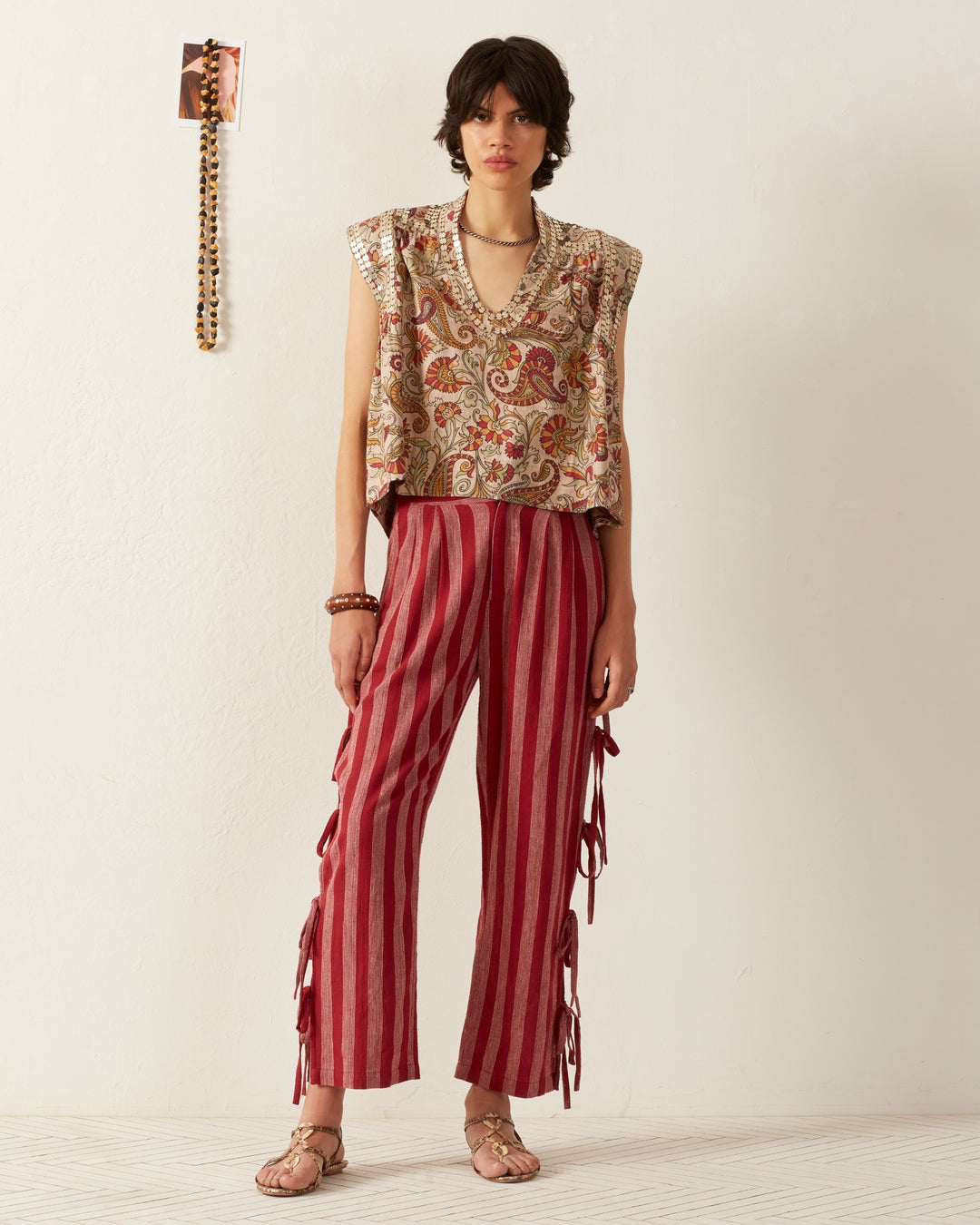 A person with short dark hair stands against a white wall, wearing a Nehru Silver Terra Top paired with red striped pants featuring ties on the sides. Sandals and some jewelry complete the look, while a decorative item hangs on the wall behind them.