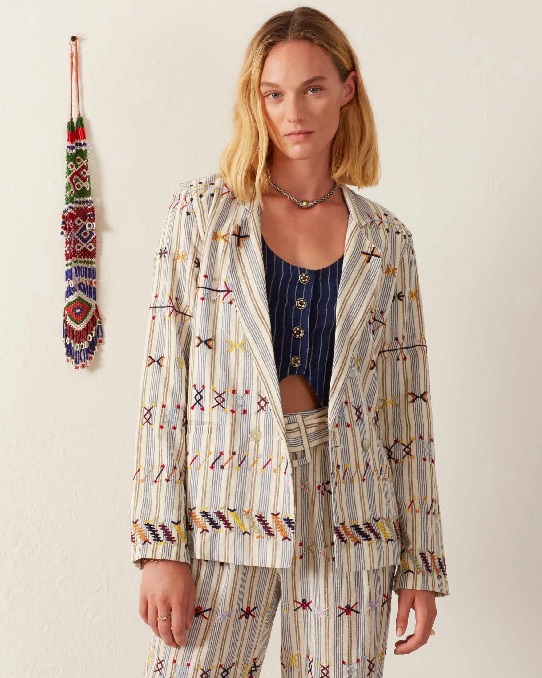 A person with shoulder-length hair wears the Elodie Arrow Linen Jacket, a beige hand-embroidered blazer featuring colorful patterns, over a blue top. Mother-of-pearl buttons enhance its elegance, while a decorative woven hanging adorns the wall in the background.