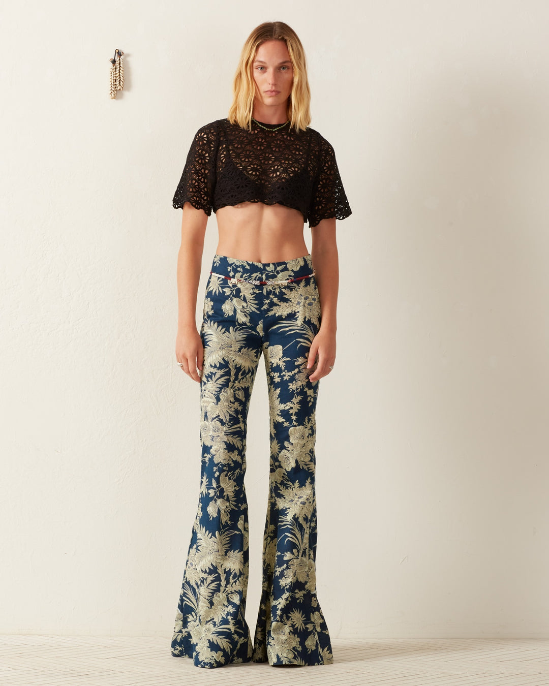 A person stands against a white wall, wearing the Nala Onyx Lace Cropped Top and blue bell-bottom pants featuring a daisy motif. They have shoulder-length hair, with a decorative item adding charm to the wall.