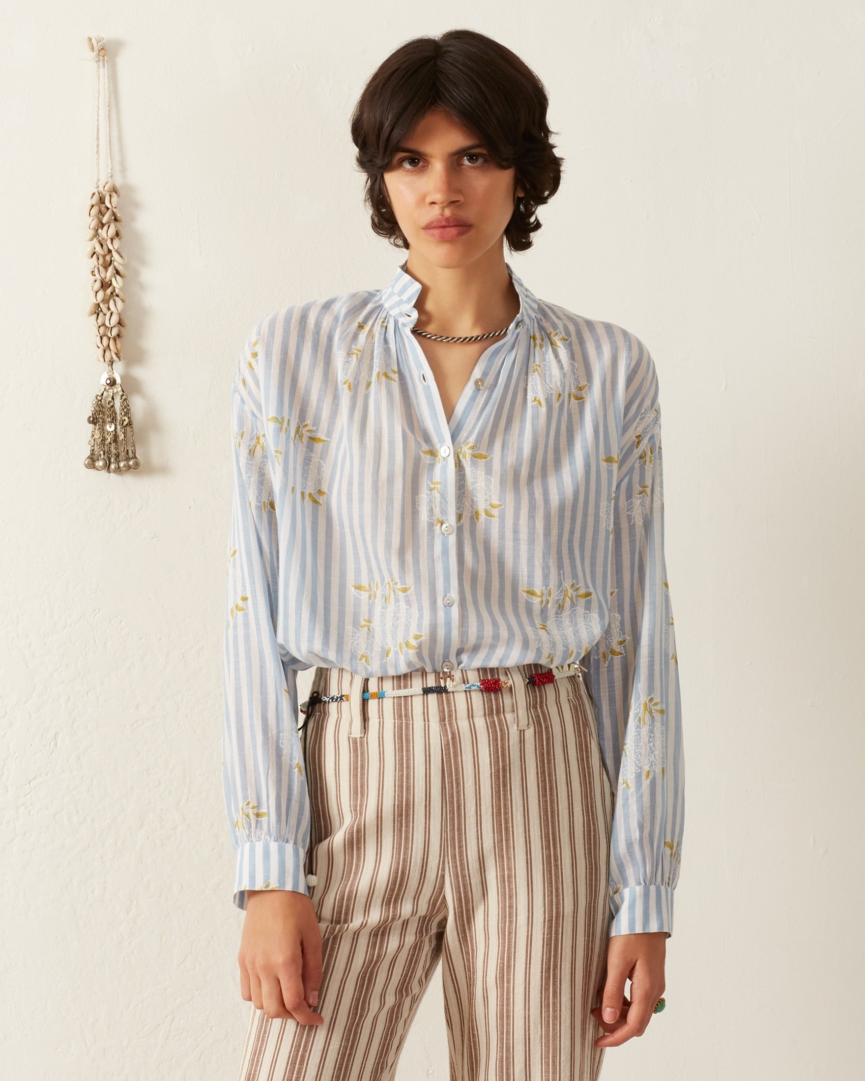 A person stands against a plain background, wearing the Kiki Magnolia Stripe Shirt, which features vintage-inspired blue and white stripes with yellow floral patterns, paired with beige and brown striped pants. The person has short dark hair and a neutral expression. A decorative tassel hangs on the wall.