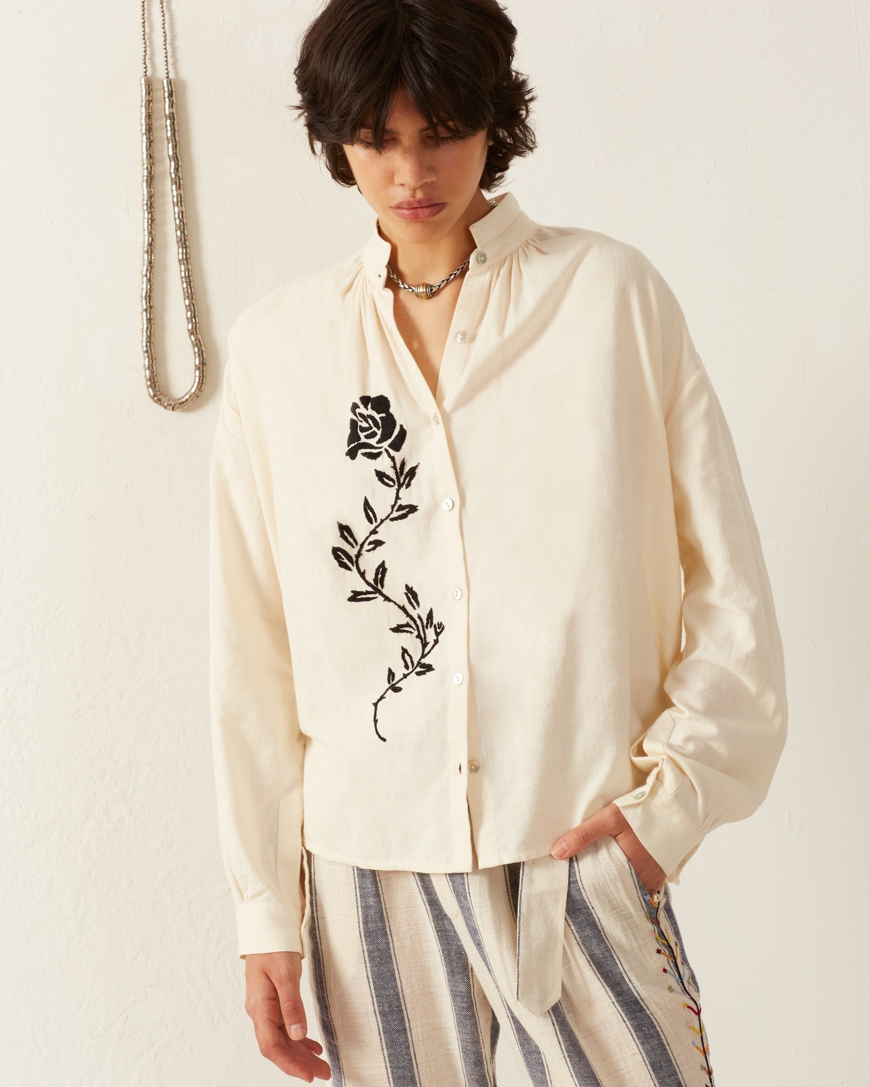 A person with dark hair wears the Kiki Noir Rose Embroidered Shirt, a cream-colored garment with a black floral design, paired with striped pants. Made from Khadi cotton, the shirt provides texture as they stand against a light-colored wall with a hanging necklace visible beside them.