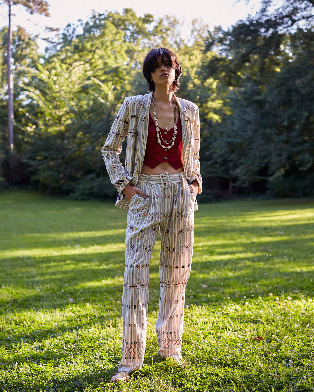 A person stands confidently on a sunlit grassy field wearing a patterned suit paired with the Colette Arrow Linen Pant and a red top. Their hands are in pockets, adorned with multiple necklaces. Tall trees and bright sunlight fill the background, adding to the serene atmosphere.