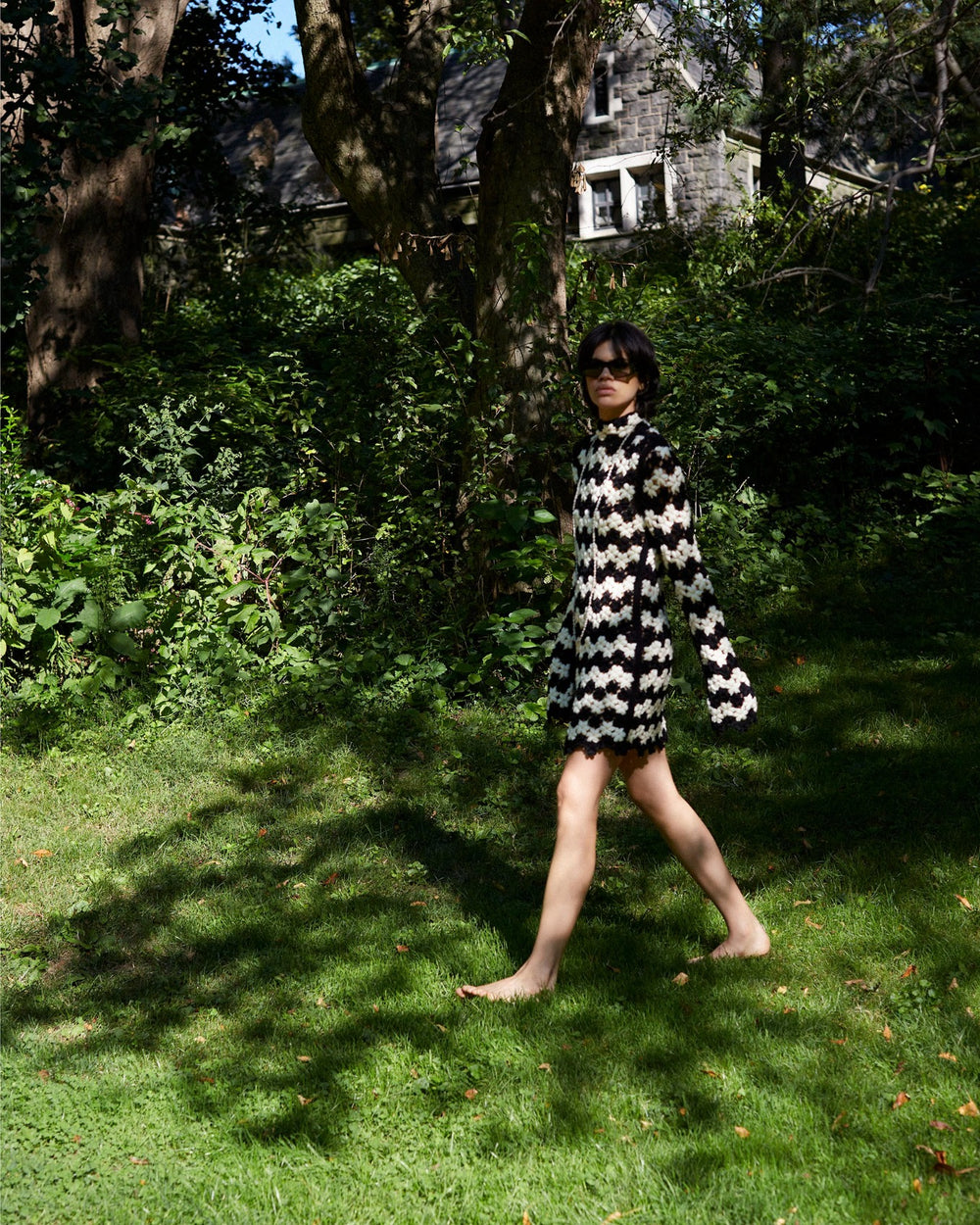 A person walks barefoot on grass in a sunlit garden, wearing the Romy Mariner Crochet Dress with a daisy motif and sunglasses. Tall trees and a house are in the background, casting shadows on the lawn, evoking a flower child style.