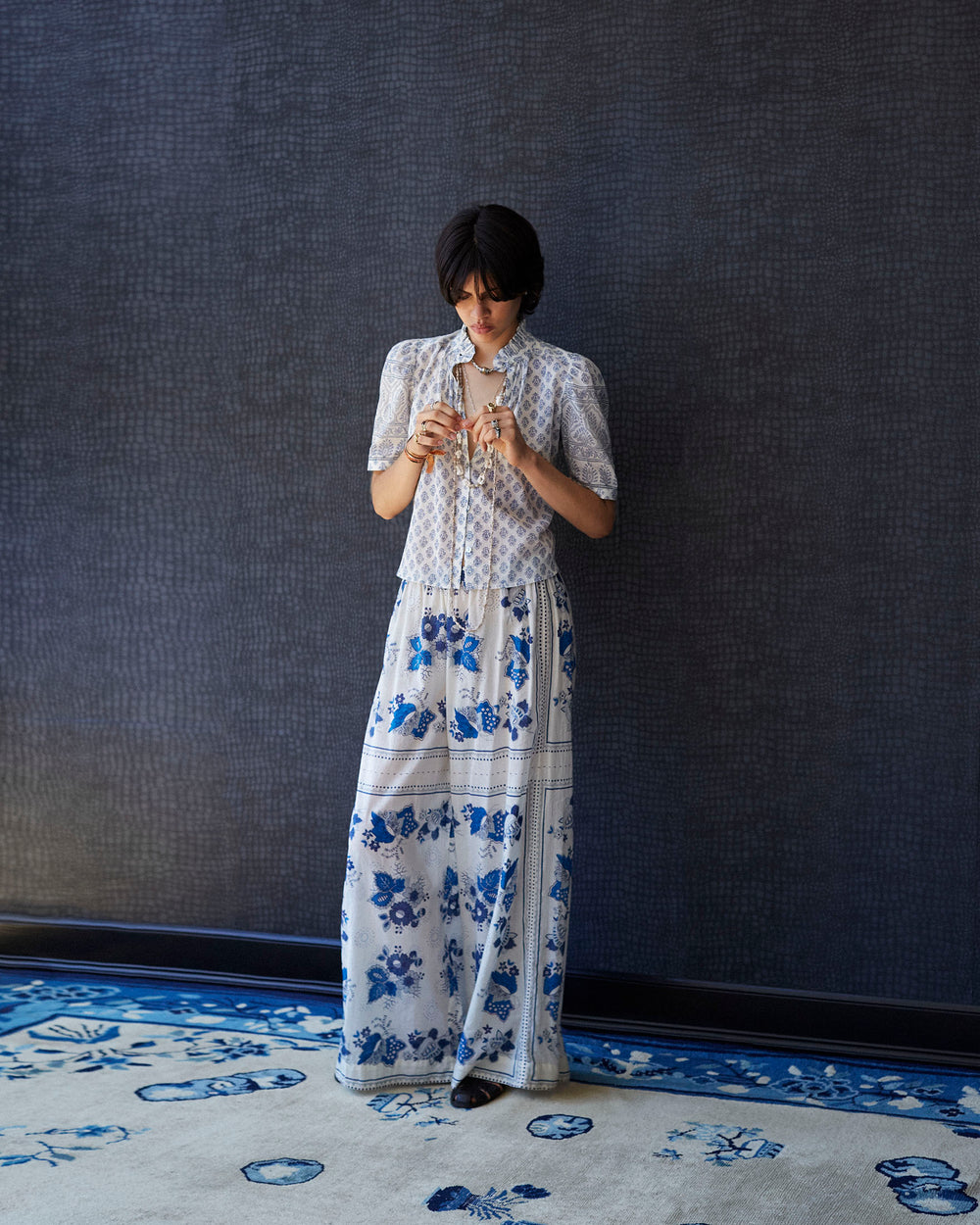 Wearing the Winnie Delft Bhutti Shirt, a person stands on a matching floral rug against a dark textured wall. They are looking down with short dark hair, seeming to adjust their glasses or jewelry.