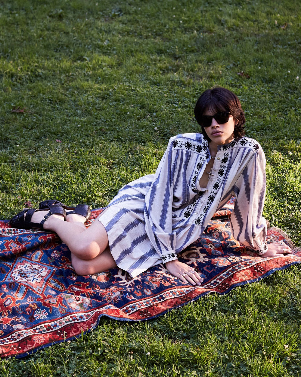 A person with short dark hair and sunglasses lounges on a patterned rug set on green grass, wearing the stunning Jolie Harbor Embroidered Dress—a hand-loomed cotton, long-sleeved striped shirt dress featuring exquisite black floral embroidery—and dark sandals.