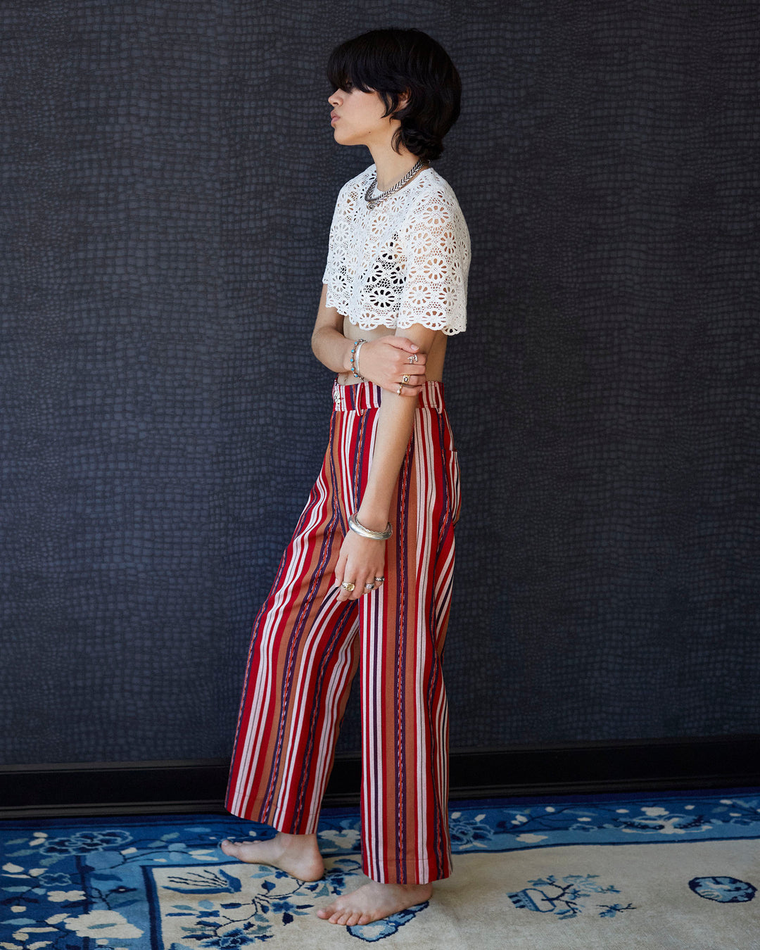 A person stands barefoot on a patterned rug against a dark, textured background. They are wearing the Nala Daisy Lace Cropped Top, a white cotton lace top adorned with an intricate daisy motif, paired with red striped pants. Their hair is short, and they are facing sideways with their arms relaxed.