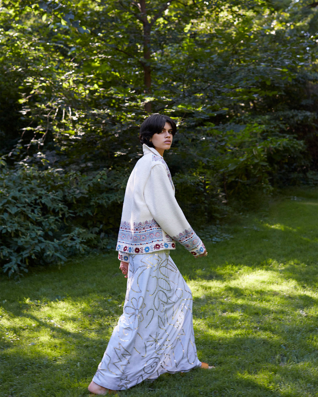 A person with short dark hair walks barefoot on grass, wearing a long, white, patterned skirt and the eye-catching Western Riviera Silk Jacket adorned with colorful embellishments. They are in a lush, green outdoor setting with sunlight filtering through the trees.