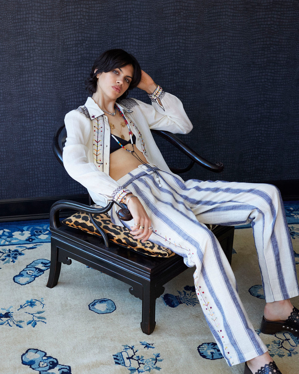 A person with short dark hair sits on a black chair, wearing the Priscilla Aurora Arrow Blouse, featuring folk-inspired patterns, along with blue-striped pants over a black bikini top. The room has a textured dark wall and a patterned blue rug on the floor.