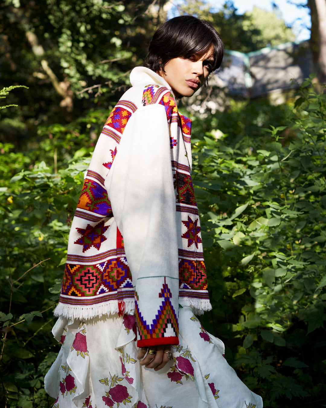 In a lush forest, someone stands wearing the Western Tiger Eye Blanket Jacket, featuring colorful, intricate geometric patterns. They pair it with a floral skirt as sunlight filters through the trees, highlighting their figure amidst the greenery.