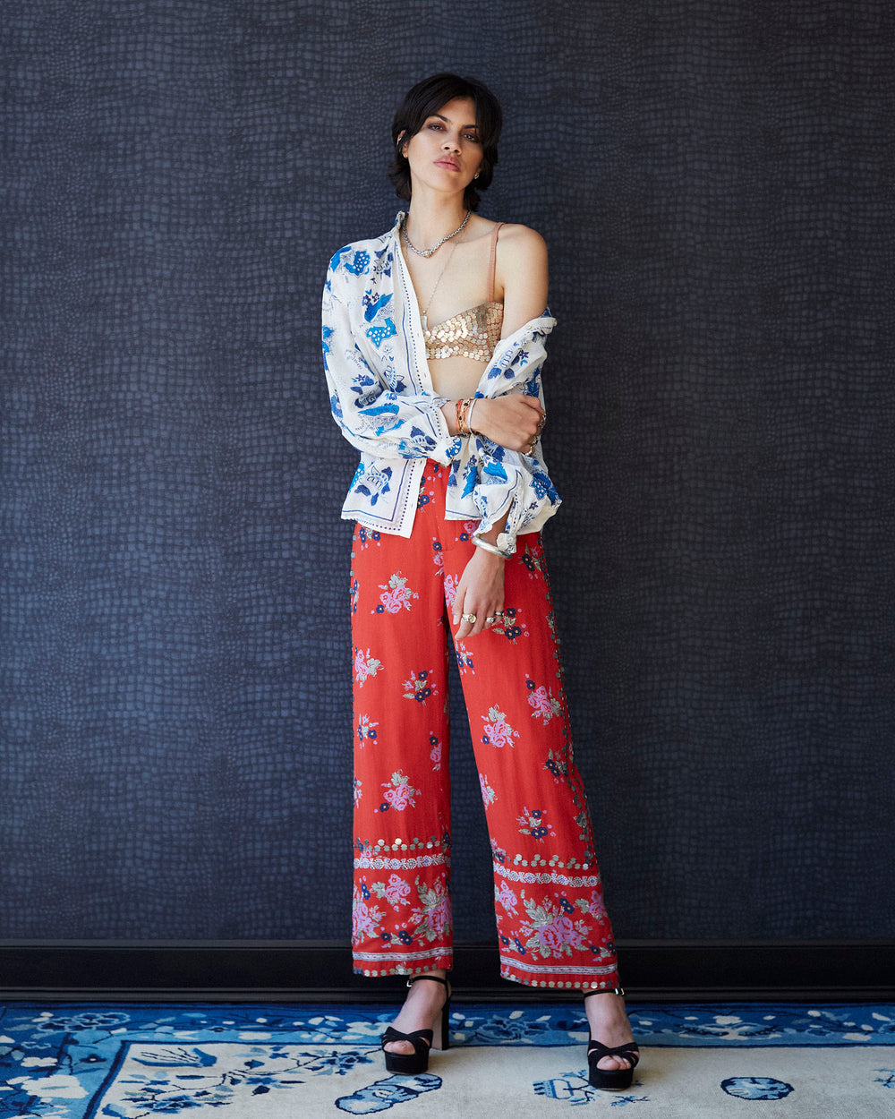 A person stands confidently against a dark textured wall, dressed in a decorative white and blue jacket over a gold top, paired with the vibrant Scout Carmine Coin Pant featuring red floral prints and complemented by black heels. The ensemble is elevated by the hand-embellished details that highlight their expressive styling.