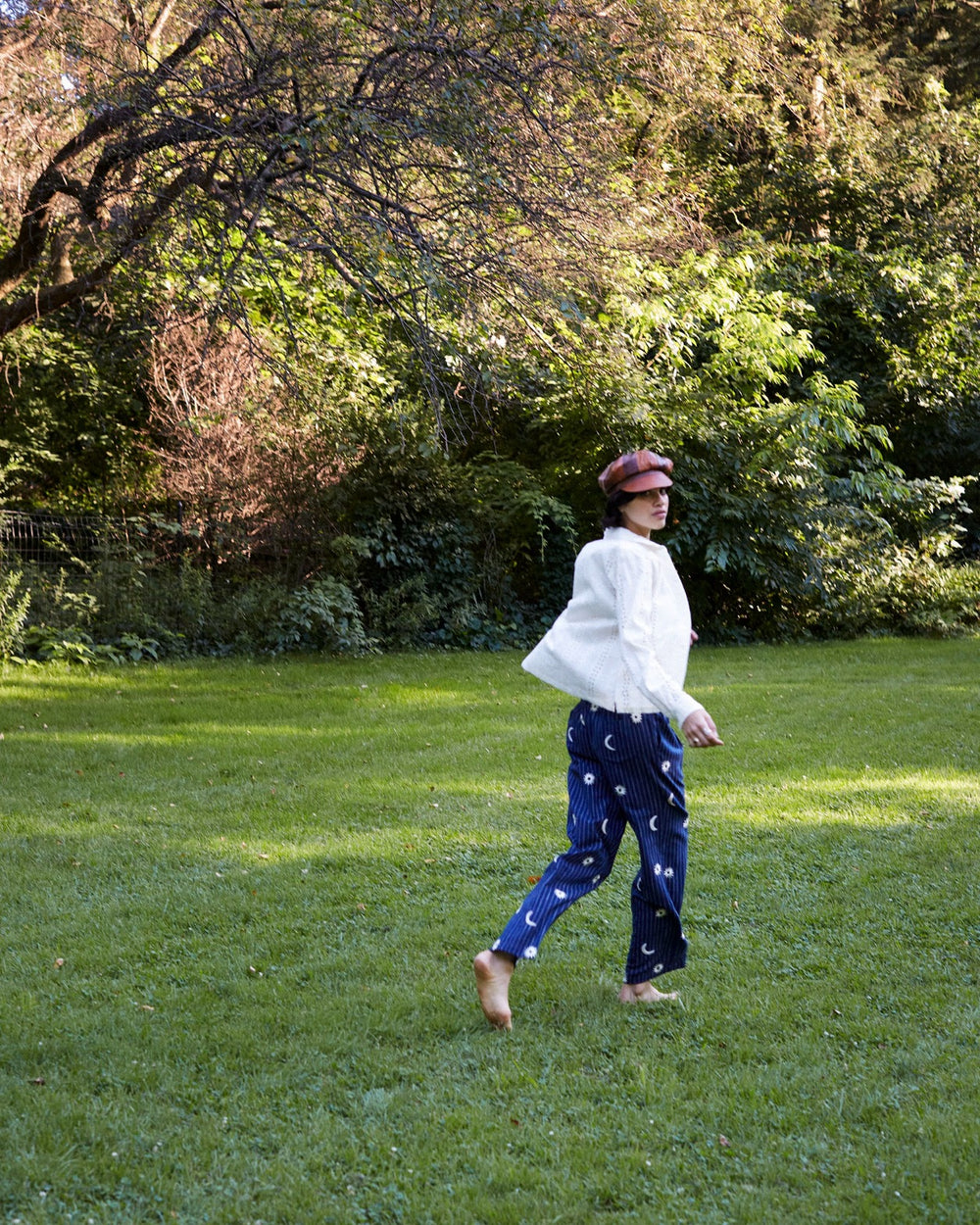 A person wearing sunglasses, a white top, and the Colette Luna Stripe Pant walks barefoot on a grassy field. The scene is bathed in natural light, with lush green trees and bushes in the background.