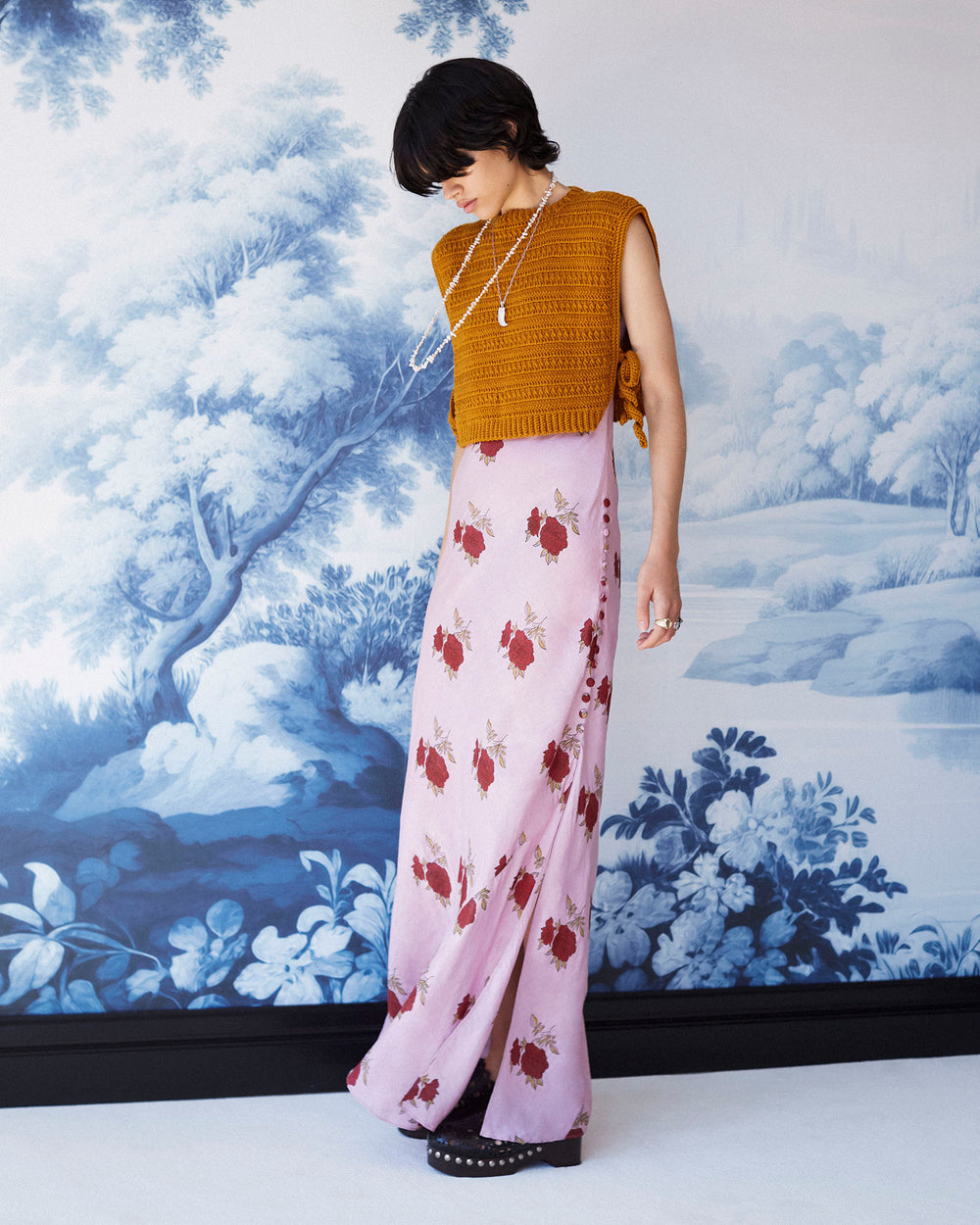 A person with short dark hair stands in front of a blue and white nature-themed wall mural. They wear an Adele Ruby Rosette Silk Dress, complemented by black shoes adorned with studs and layered necklaces.