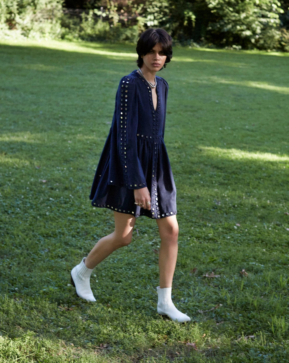 A person wearing the Silvie Midnight Mirror Dress, featuring hand-embroidered eyelet details, and white boots walks across a grassy field under the sunlight. Trees and bushes are visible in the background.