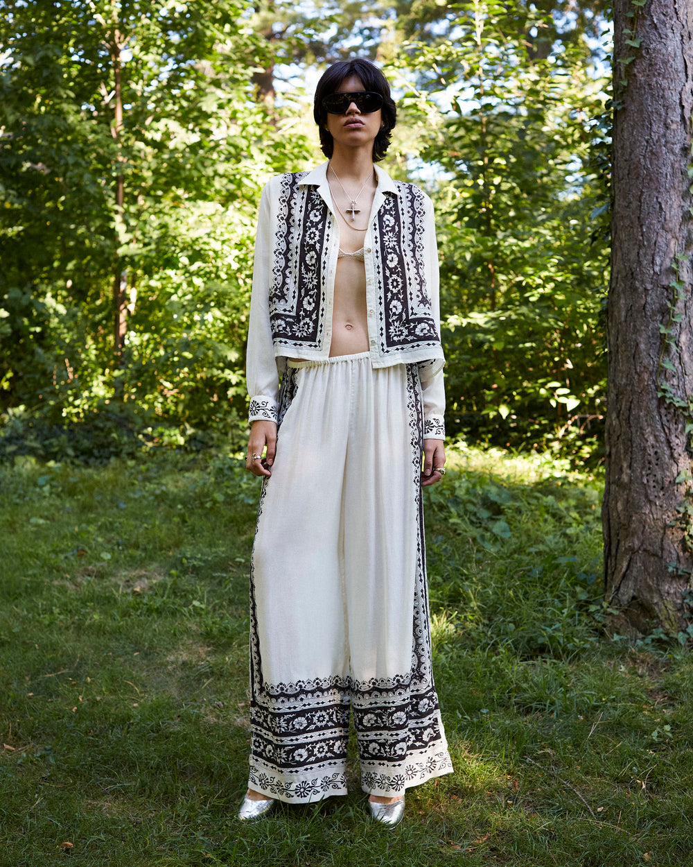 In a forest setting, an individual is dressed in the Margie Noir Riviera Pant ensemble, which includes a cream outfit with a black border print motif. The attire consists of a long-sleeved top paired with wide-leg silhouette pants. They have short dark hair and wear sunglasses, while silver shoes add the finishing touch to this Tencel creation.