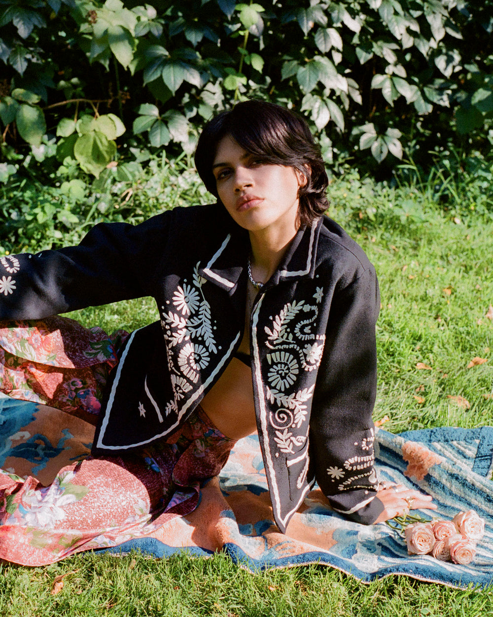 A person with dark hair sits on a colorful blanket outdoors, wearing the Francis Onyx Snail and Snake Jacket, known for its intricate floral embroidery and black silk design, paired with patterned pants. Green foliage is visible in the background, and sunlight casts a warm glow, highlighting the artisan garment's exquisite details.