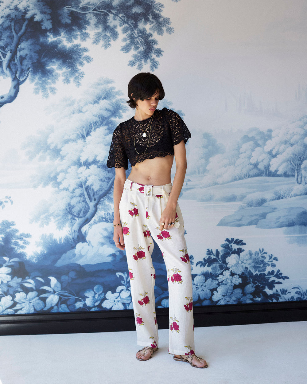 A person stands in front of a backdrop showcasing blue and white tree-covered hills, wearing sunglasses, a black lace crop top, and the Colette Cerise Rosette Pant featuring a pink rose print. They complete their look with layered necklaces, short hair, and sandals.