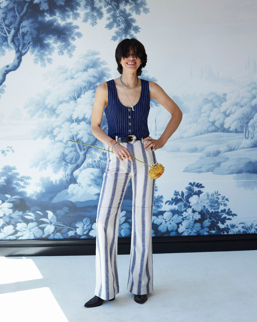 A person stands smiling in front of a scenic blue and white mural, exuding a nautical style. They wear a sleeveless blue top and striking Charlie Harbor Stripe Pants while holding a golden flower. Their hair partially covers their face, adding an air of mystery.