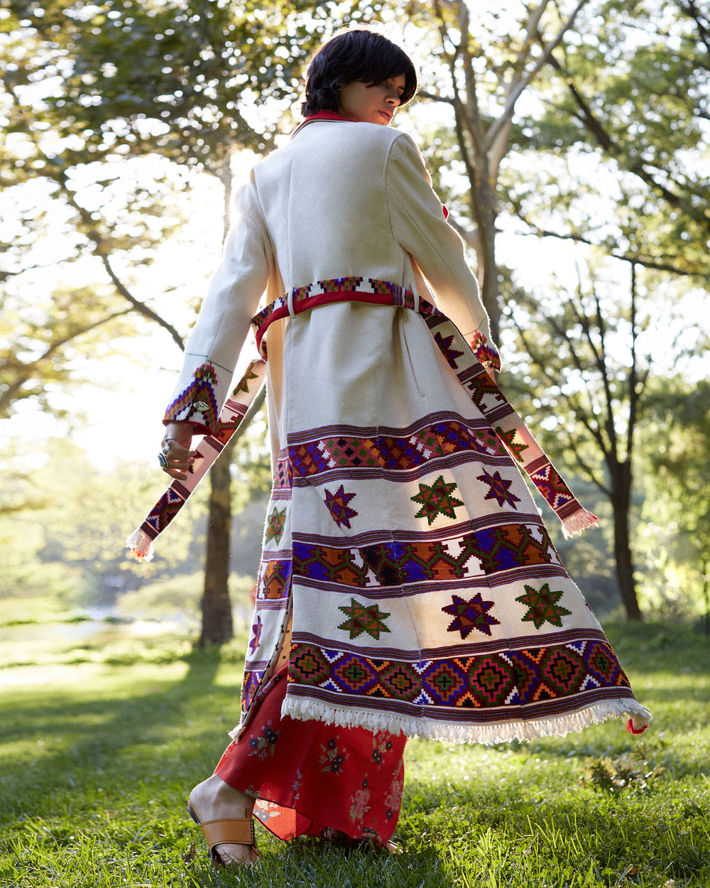 A person in the Penelope Tiger Eye Blanket Trench, showcasing intricate Southwest-style patterns, stands outdoors in a sunlit park. They hold the matching belt of the trench and wear pants adorned with red floral patterns. Trees and grass provide a serene natural backdrop, enhancing the vibrancy of the ensemble.