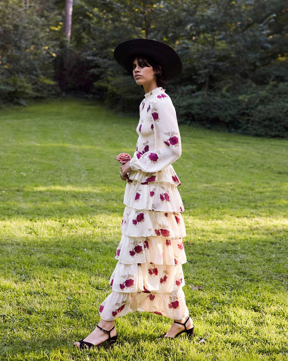 A person in a Shelby Cerise Rosette Dress, featuring layers and red flower patterns, stands on a lush green lawn, wearing a black wide-brim hat. The floral dress exudes elegance amidst the backdrop of towering trees.