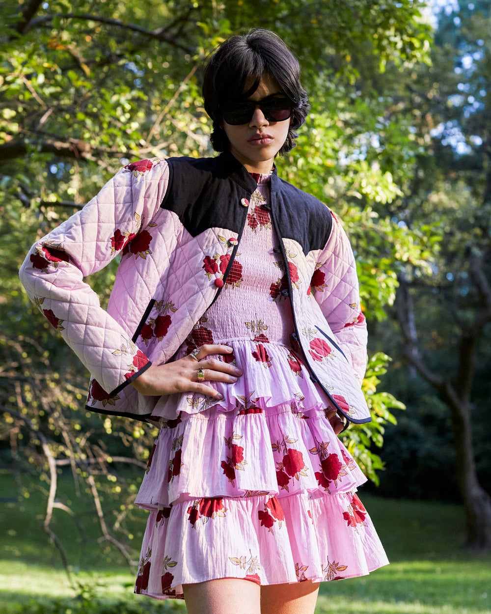 A person outdoors wearing a quilted jacket and the Cabana Ruby Rosette Short Dress, which features a layered pink floral design with red patterns and a smocked bodice, accessorized with sunglasses. There are trees and greenery in the background, indicating a park setting.