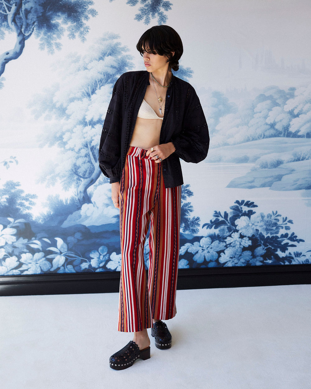 A person stands in front of a blue and white scenic mural, showcasing effortless style with their cream bralette, black open jacket, and Paige Canyon Stripe Pant. They complete the outfit with black clogs and short dark hair.