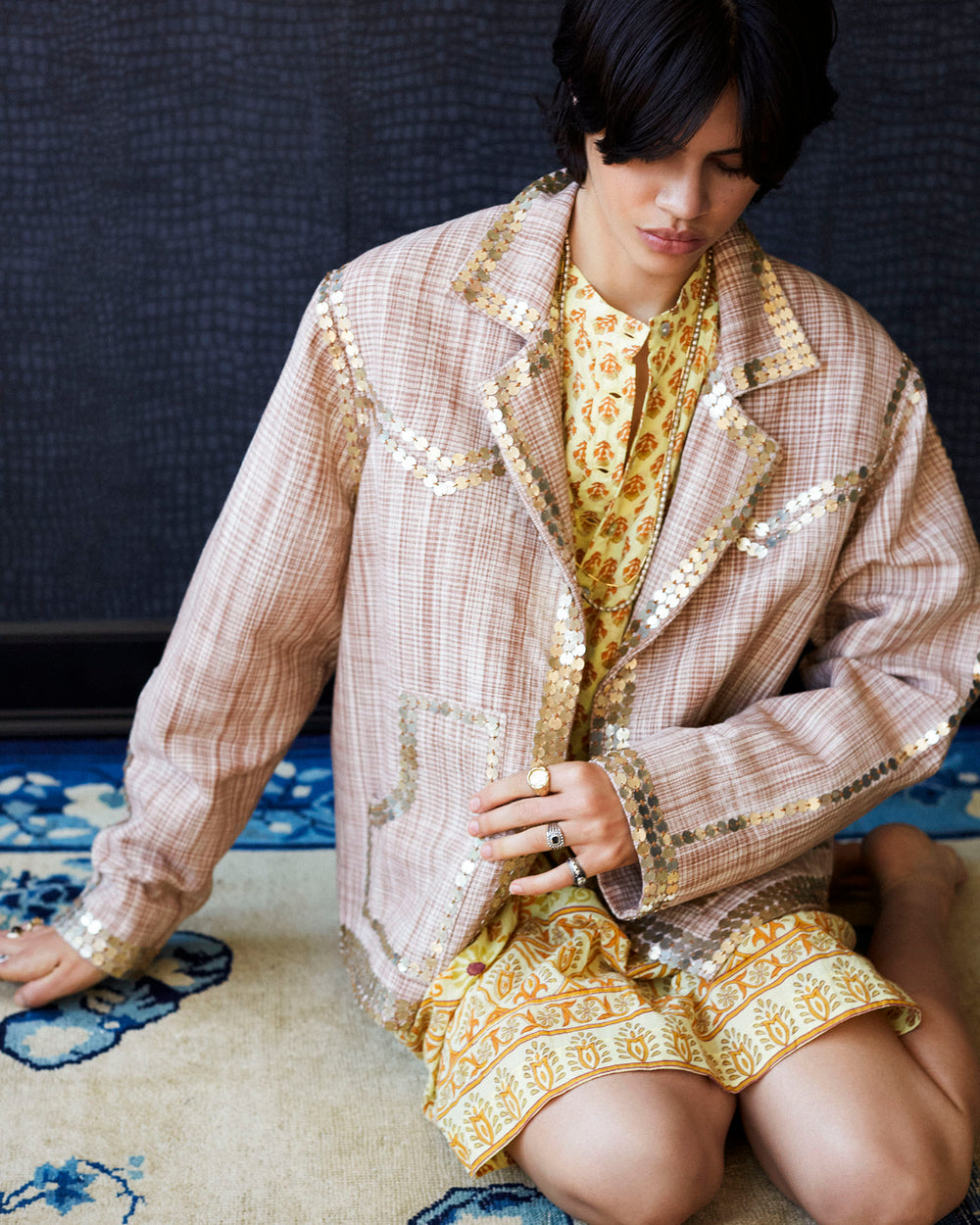 A person with short hair sits on a patterned rug, wearing the Western Sand Coin Jacket, crafted from Khadi cotton and adorned with hand-embellished metallic details. They pair it over a yellow floral dress while accentuating their look with several rings as they hold the jacket closed, casting a thoughtful gaze downward.