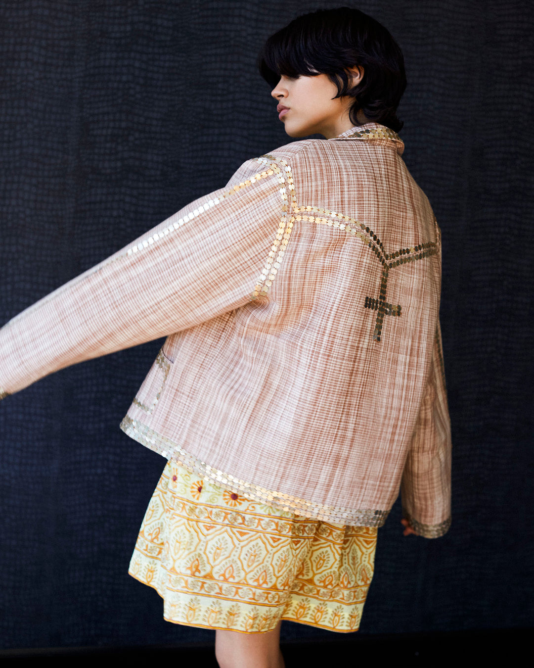 A person with short, dark hair faces sideways, wearing the Western Sand Coin Jacket featuring metallic details and embroidered patterns, paired with a yellow patterned skirt crafted from Khadi cotton. The background is dark and textured.