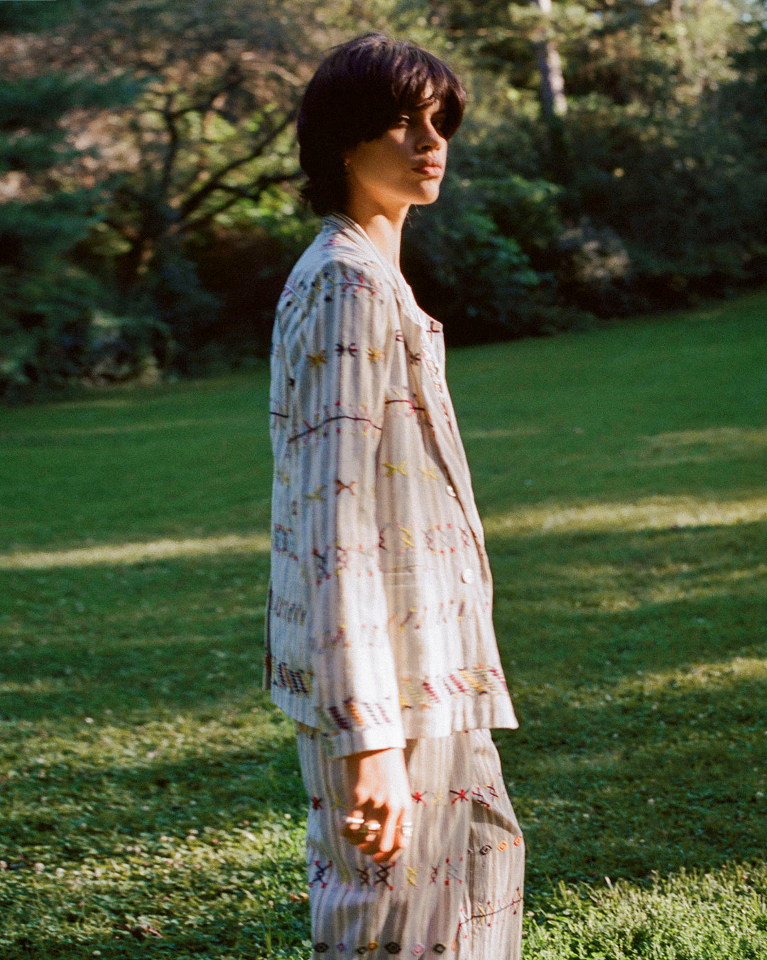 A person stands outdoors in a garden wearing the Elodie Arrow Linen Jacket, which features mother-of-pearl buttons and a matching patterned design. The light-colored jacket boasts multicolored embroidery, and sunlight filters through the trees, casting a soft, natural glow on the scene.