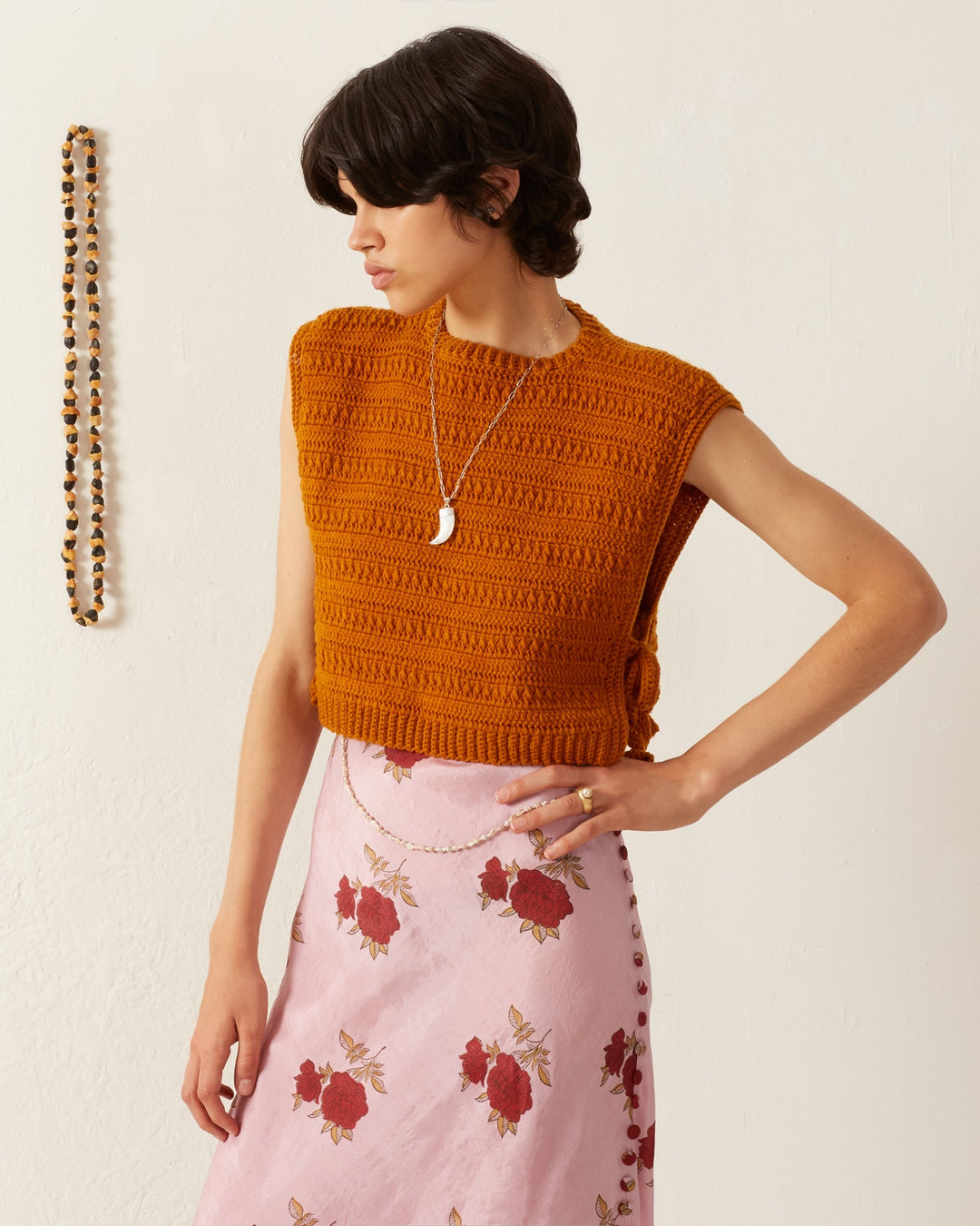 A person with short dark hair wearing the Oona Raffia Crochet Top, a vintage-inspired sleeveless orange knit top, and a pink floral skirt stands against a plain light background. A necklace with a tooth pendant and a beaded wall decoration are visible.