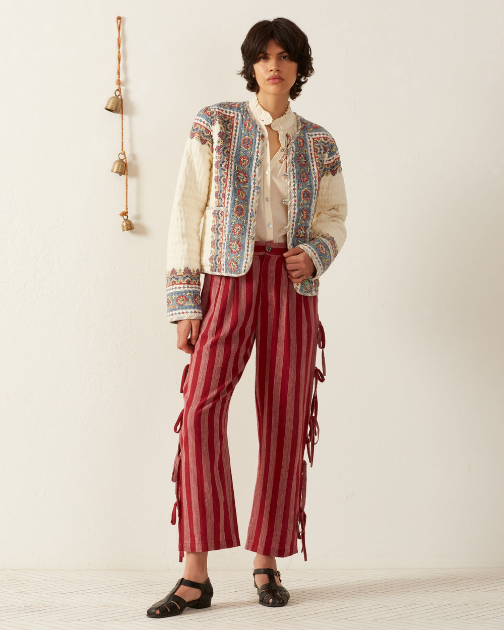 A person is wearing the Pasha Riviera Jacket, featuring a block-printed cream and blue pattern, over a white blouse. They've paired it with red and white striped pants that have side ties. Against a plain light-colored wall with hanging bells, they complete the look with black sandals.